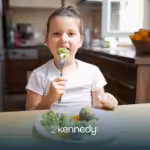 A girl with autism sitting at a table, eating a piece of broccoli