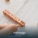 A toddler sitting on a floor, pointing at wooden blocks that spells the word 'autism'